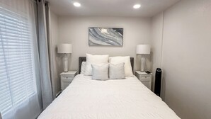 A neatly made bed with white linens and pillows, two bedside tables with lamps, a wall-mounted artwork, and a window with blinds. - Prime Spot! Walk to Ski, Shops, and Main Street! (Park City)