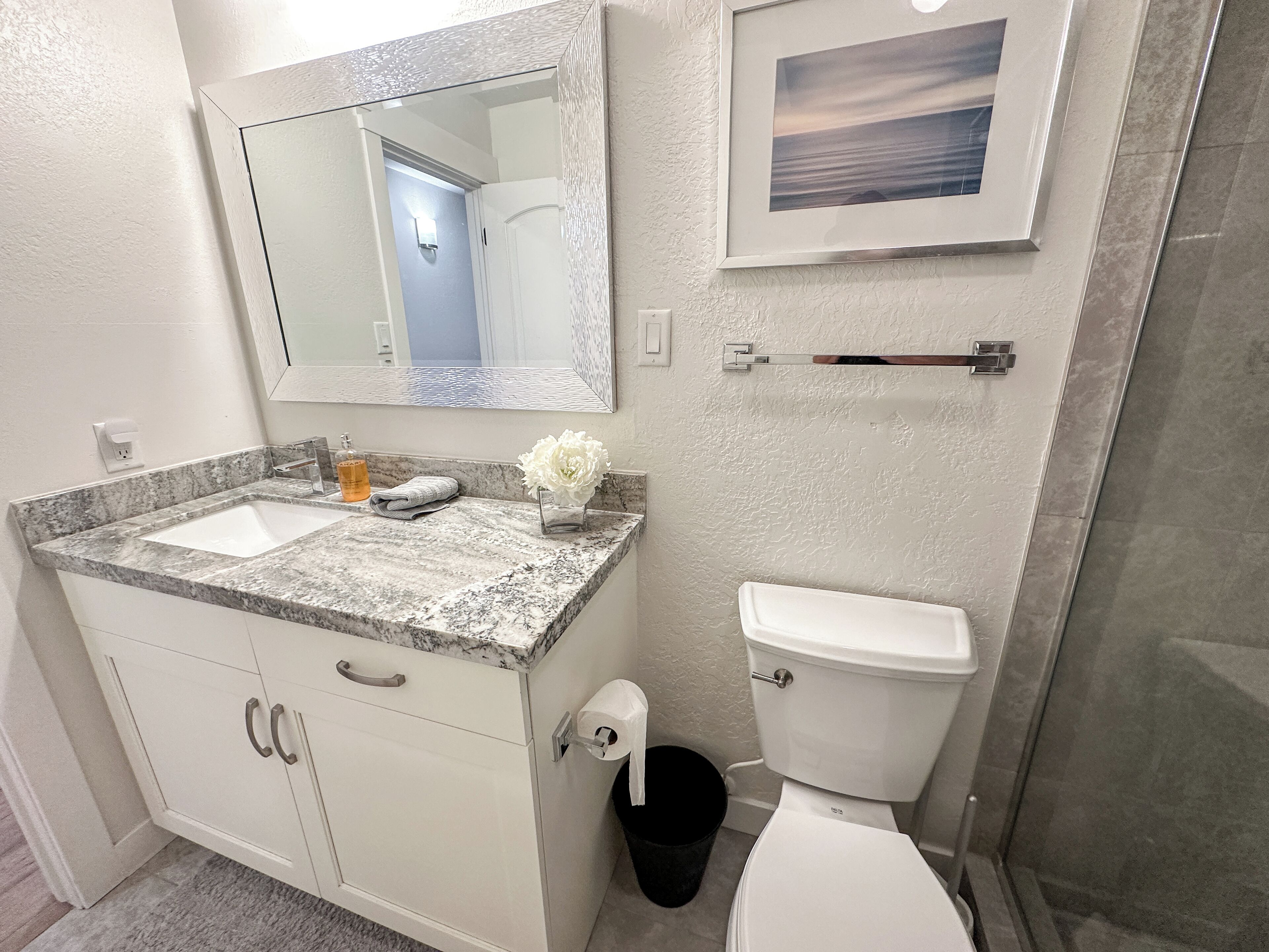 A bathroom with two sinks, a toilet, a trash can, a towel, and a framed picture on the wall.