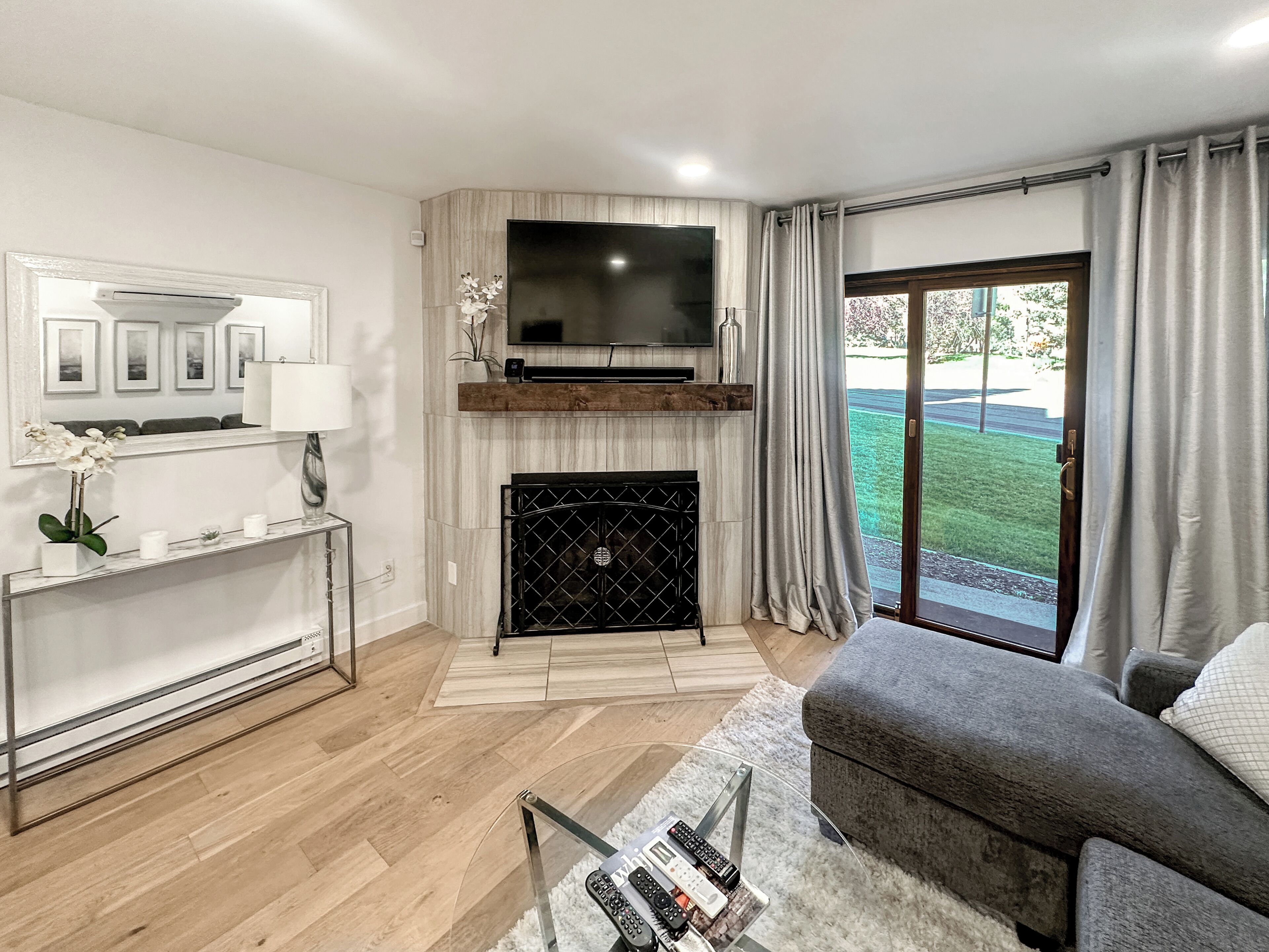 Un salon doté d’une cheminée, d’un téléviseur à écran plat fixé au-dessus, d’un canapé, d’une table basse avec des télécommandes, et offrant une vue sur un espace verdoyant grâce à une porte-fenêtre coulissante.