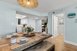 A dining area with a wooden table set for four, a bench, and a wall clock. - Oceania Beach Club 103 (New Smyrna Beach)