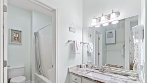 A bathroom with a white toilet, a shower curtain, a sink with a marble countertop, a large mirror, and a hairdryer on the wall. - Royal Poste Unit 3203 (Sunset Beach)