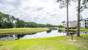 A tranquil riverside view with a grassy area, a small pond, and a building with multiple floors in the background. - Sandpiper 2A (Sunset Beach)
