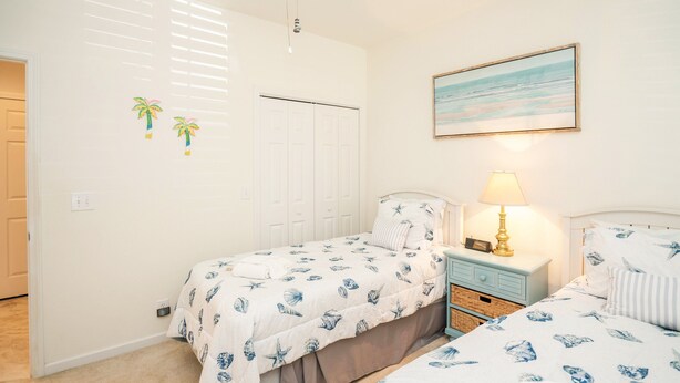 A bedroom with two beds, a nightstand with a lamp, a framed picture on the wall, and a closet. - Sandpiper 2A (Sunset Beach)