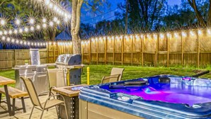 An outdoor hot tub with a wooden deck, a grill, and string lights. - Sandhill Flat (Gary)