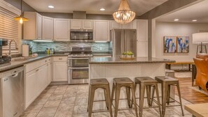 A modern kitchen with white cabinets, stainless steel appliances, a large island with bar stools, and a chandelier. - Sandhill Flat (Gary)