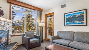 A cozy living room with a fireplace, a sofa, a chair, and a large window offering a mountain view. - Serene 1BR Rockies Retreat w/ BBQ, Gym & Hot Tub (Canmore)