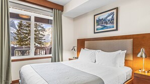 A hotel room with a large bed, a wooden headboard, a nightstand with a lamp, a window with curtains, and a framed picture on the wall. - Serene 1BR Rockies Retreat w/ BBQ, Gym & Hot Tub (Canmore)
