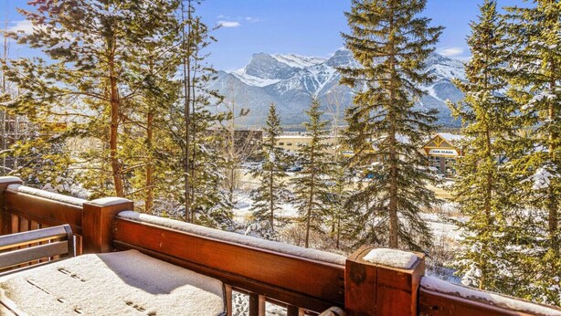 A wooden deck with a view of snow-covered mountains and a car rental sign. - Serene 1BR Rockies Retreat w/ BBQ, Gym & Hot Tub (Canmore)