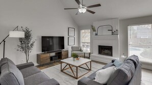 A modern living room with a flat-screen TV, grey sofas, a coffee table, a fireplace, and a ceiling fan. - The Prestige Poolside Escape - 5BR w Cinema & Gaming (DeSoto)