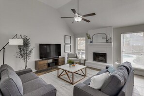 A modern living room with a flat-screen TV, grey sofas, a coffee table, a fireplace, and a ceiling fan.