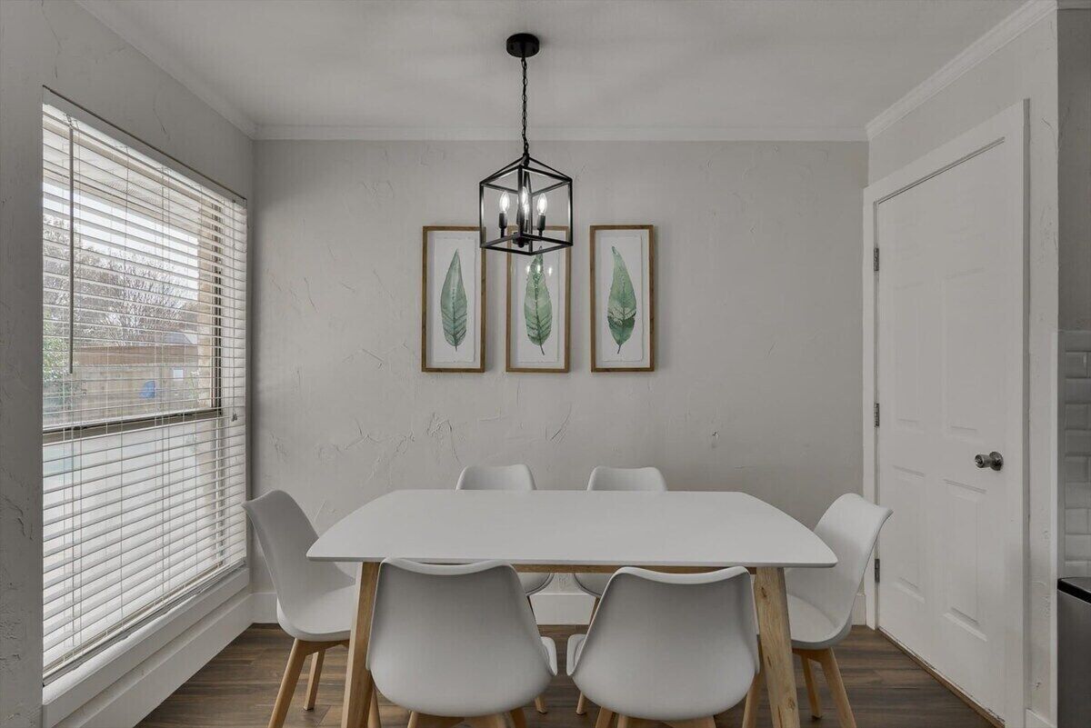 A dining area with a white table, six chairs, a hanging light fixture, and framed leaf artwork on the wall.