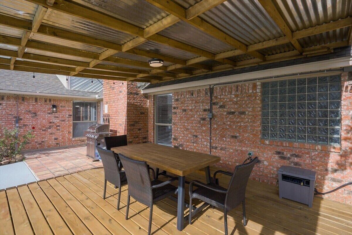 Patio esterno con tavolo e sedie in legno, un barbecue e un muro di mattoni con finestre.