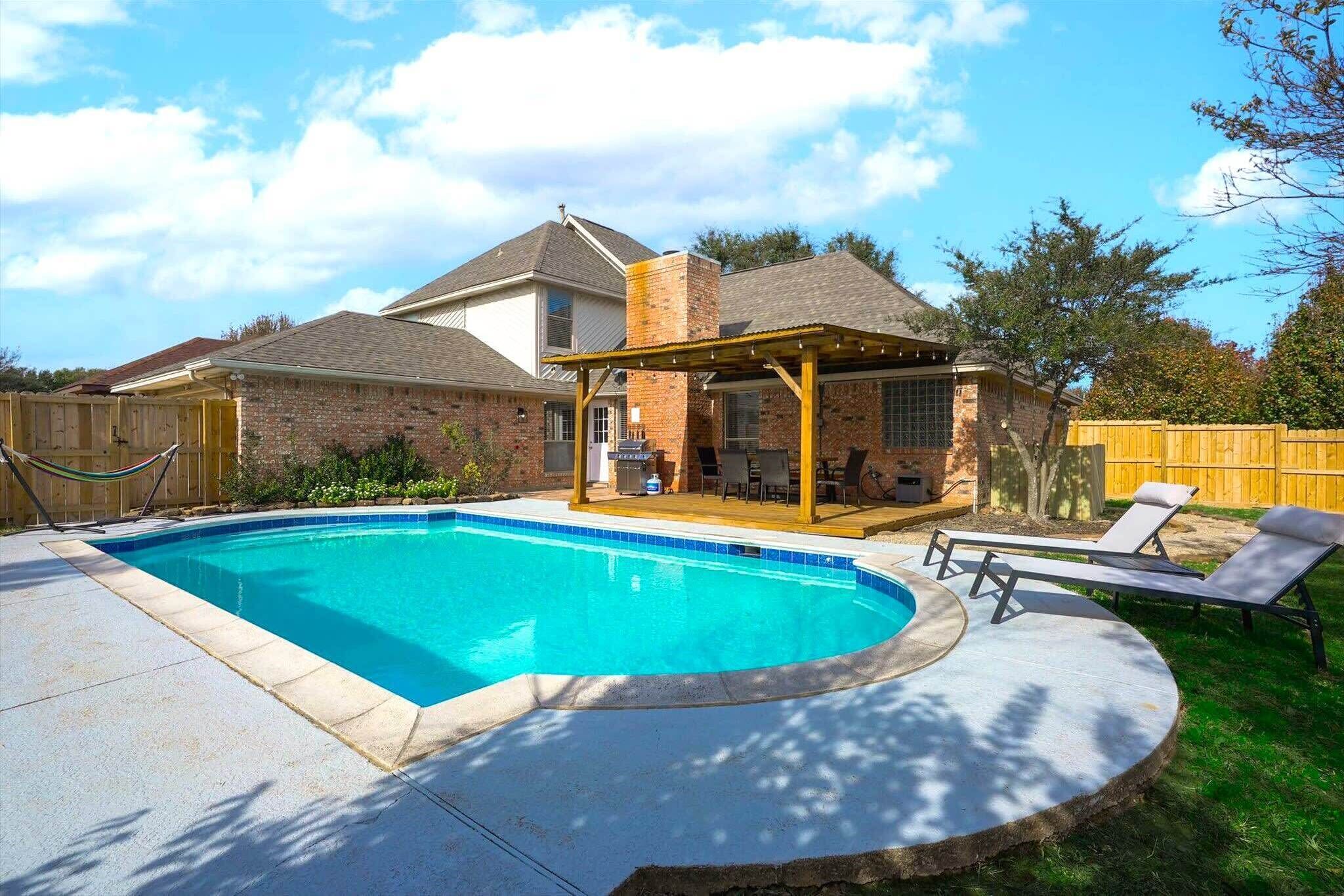 A backyard with a swimming pool, a covered patio, and lounge chairs.