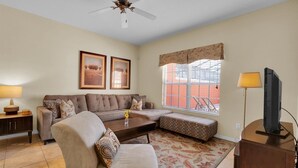 A living room with a sofa, armchair, and a television on a stand. There is a ceiling fan, a window with blinds, and a patterned rug. - Cpr8972 (Kissimmee)