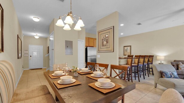 A dining area with a wooden table set for four, surrounded by chairs. There is a kitchen in the background with wooden cabinets and a refrigerator. - Cpr8972 (Kissimmee)