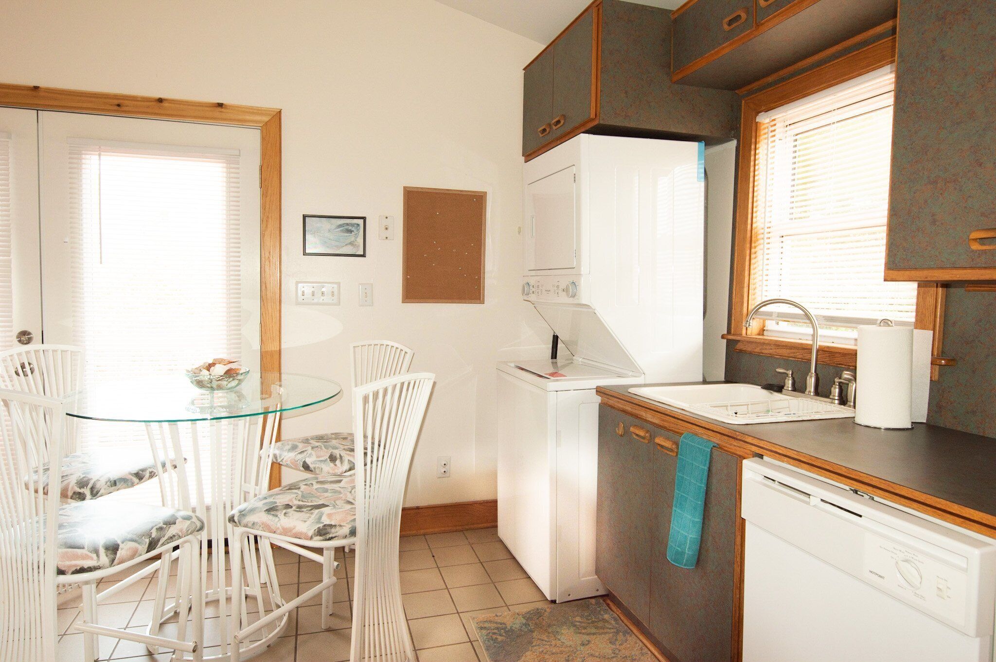 A compact kitchen with a glass dining table, white chairs, a white refrigerator, a dishwasher, and wooden cabinets.
