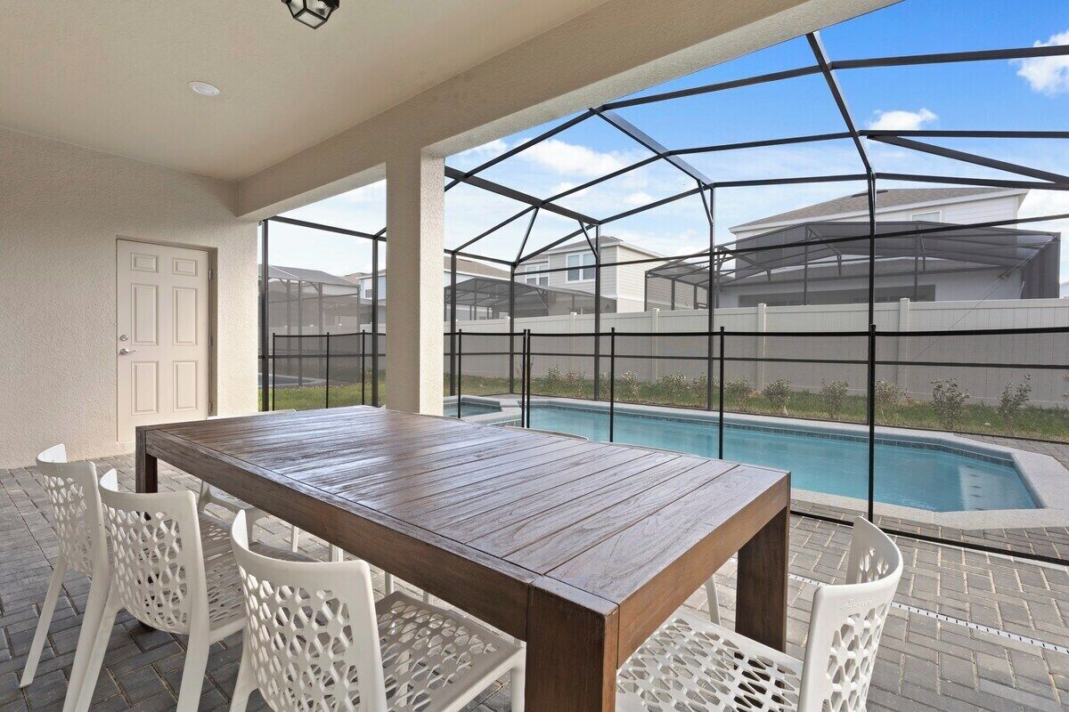 A patio area with a wooden table and white chairs, overlooking a swimming pool and a residential neighborhood.
