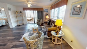 A living room with a sofa, armchair, and glass coffee table. There is a window with blinds, a ceiling fan, and a lamp on a side table. - River Creek 1 Unit 304 (Sunset Beach)
