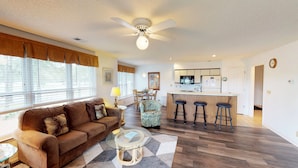 A living room with a brown sofa, a glass coffee table, and a patterned armchair. There is a kitchen area with a bar and stools, and a dining table with chairs. - River Creek 1 Unit 304 (Sunset Beach)