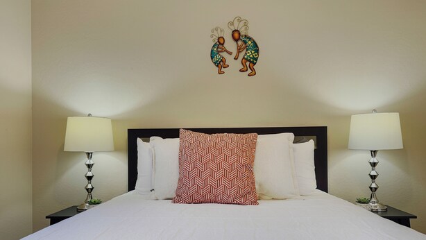 A neatly made bed with white linens and a red patterned pillow, flanked by two bedside tables with lamps, in a room with a plain wall and a decorative wall hanging above the bed. - Southern #3116 Mesa (Mesa)