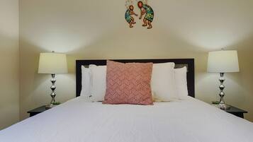 A neatly made bed with white linens and a red patterned pillow, flanked by two bedside tables with lamps, in a room with a plain wall and a decorative wall hanging above the bed.