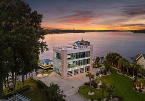 A modern building with a glass facade, a covered outdoor area, and a view of a lake at sunset. - The Glass Mansion (Terrell)