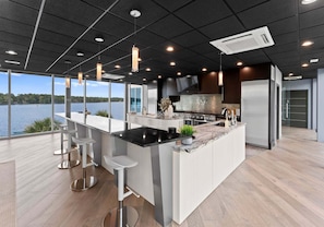 A modern kitchen with a large island, bar stools, and a view of a lake. - The Glass Mansion (Terrell)
