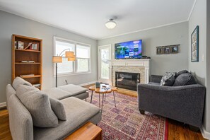 A living room with a grey sofa, a grey armchair, a wooden bookshelf, a floor lamp, a flat-screen TV, and a fireplace. - Plan A Trip To Georgia Aquarium In This Comfy 3BR (Hapeville)