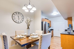 Un comedor con una mesa de madera dispuesta para cuatro personas, un reloj de pared grande y un jarrón con flores secas.