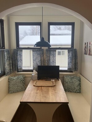 Ein gemütliches Zimmer mit Schreibtisch aus Holz, zwei Fenstern mit Vorhängen, einer Hängelampe und Blick auf eine verschneite Landschaft.