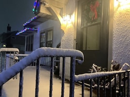 Un área exterior cubierta de nieve con una baranda de metal, la entrada de un edificio con una pared iluminada y una ventana con decoración roja.