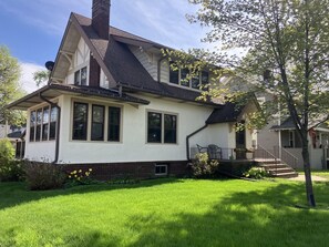 A two-story house with a gabled roof, a front porch, and a small front yard with a tree and flowers. - Sax Zim Bog Lodge Hibbing Downtown Home - Located Downtown Hibbing, (Galveston)