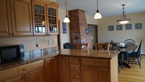A kitchen with wooden cabinets, a brick fireplace, pendant lights, and a dining area with chairs and a table. - Short Sands View Y770-g (York Beach)