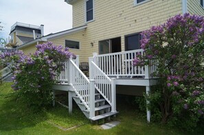 Ein zweistöckiges Gebäude mit einer weißen Treppe, die zu einem Balkon führt, umgeben von üppigem Grün und violetten Blumen.
