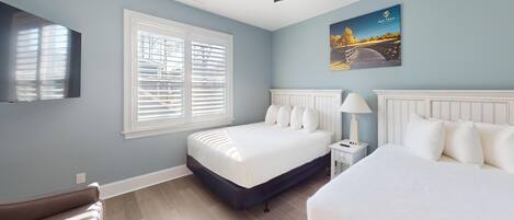 A bedroom with two beds, a window with blinds, a bedside table with a lamp, and a wall-mounted TV.