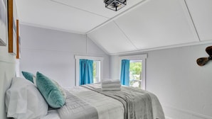 Un dormitorio con una cama, dos ventanas con cortinas azules, un ventilador de techo y una lámpara fijada a la pared.