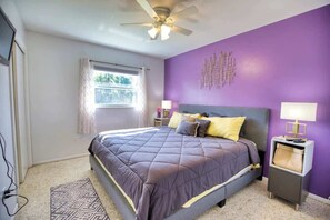 Un dormitorio con cama, ventilador de techo, ventana con cortinas y pared morada.