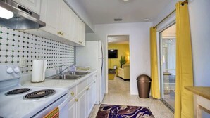 A compact kitchen with white cabinets, a stove, and a sink. A patterned rug is on the floor, and a trash can is visible. - Stylish Retro Vibes Private BBQ & Beach Access (Sarasota)