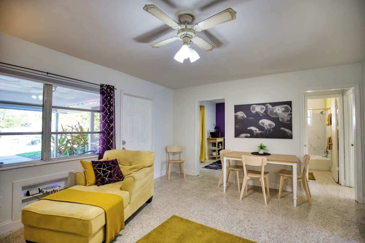 A compact living space with a yellow sofa, a dining table with chairs, a ceiling fan, and a large window.