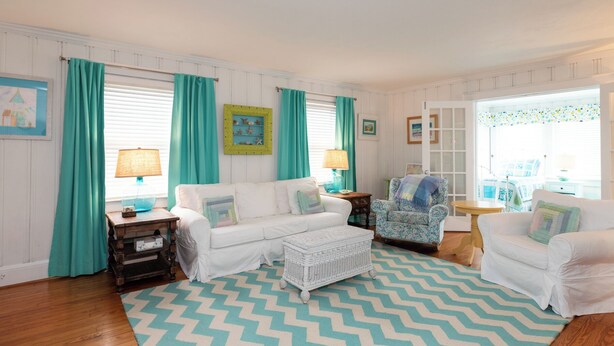 Un salon chaleureux avec un canapé blanc, un tapis à motifs, des rideaux turquoise et une petite table d’appoint surmontée d’une lampe.