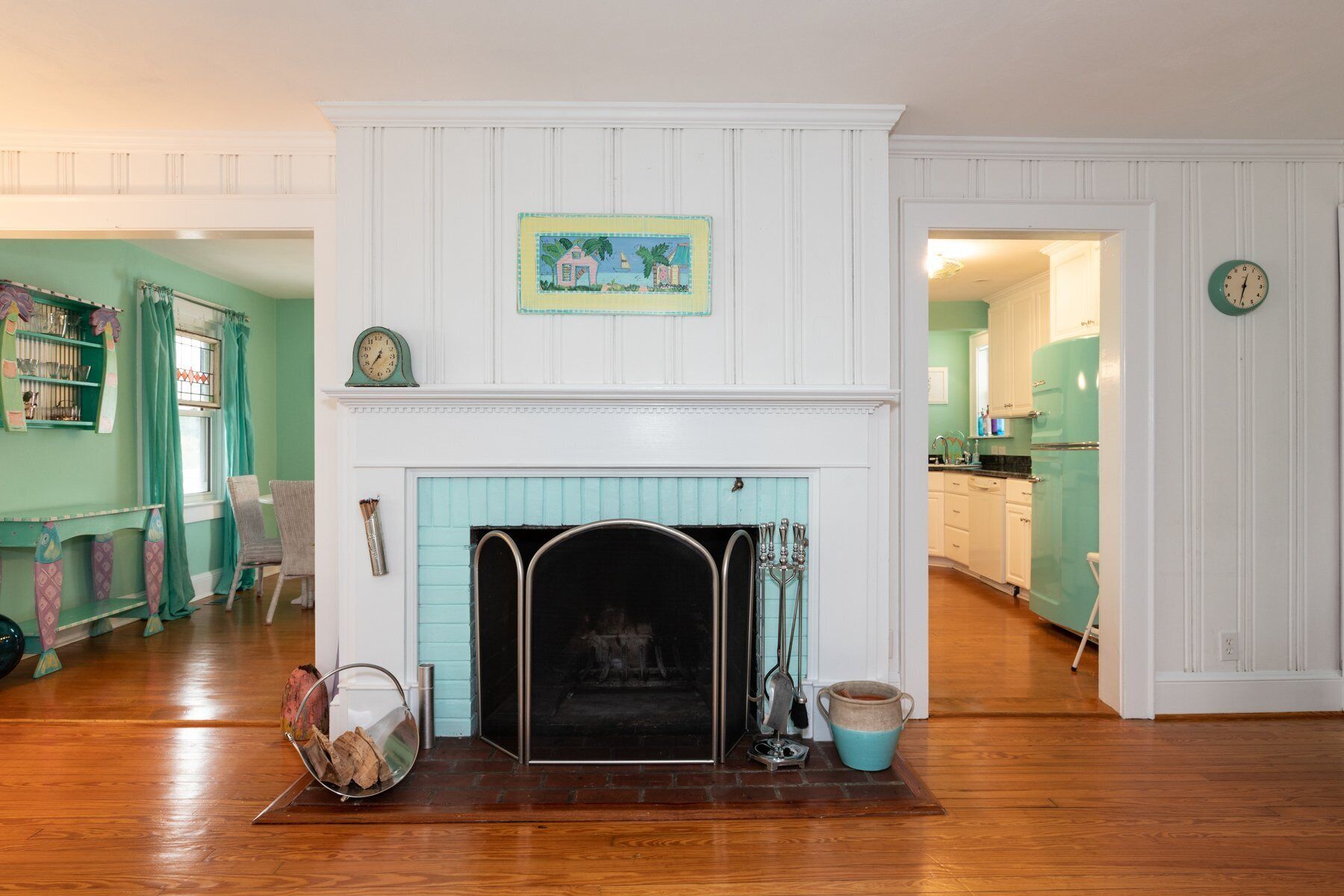 A room with a fireplace, a clock on the wall, and a kitchen visible through an open door.