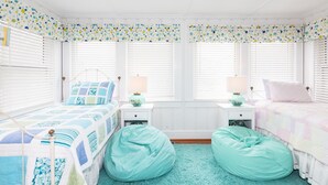 Une chambre avec deux lits, une fenêtre avec des stores, une lampe sur chaque table de chevet et un fauteuil en forme de poire bleu.