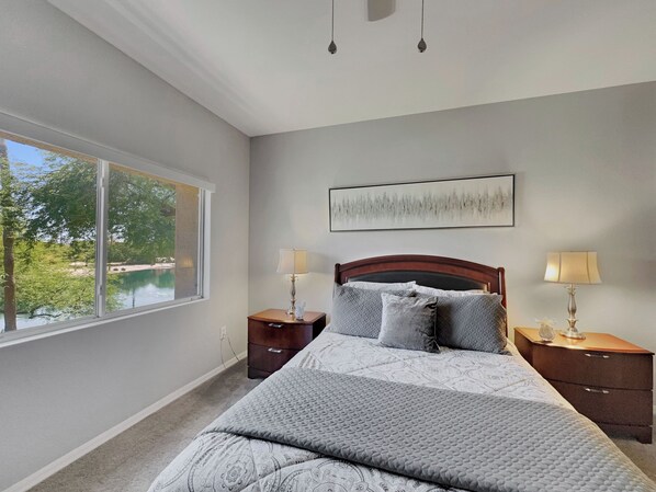 A bedroom with a large window, a bed with a quilt, two bedside tables with lamps, and a painting on the wall. - Superstition Springs Mesa (Mesa)