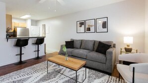 A modern living room with a grey sofa, two black bar stools, a wooden coffee table, and a ceiling fan. - The Cozy Corner Near Downtown (Athens)