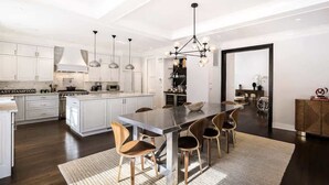 A modern kitchen with white cabinetry, a central island, and a dining area with wooden chairs. - Bull Path Manor by Rove - 4BR Family Home With Pool & Hot Tub (East Hampton)