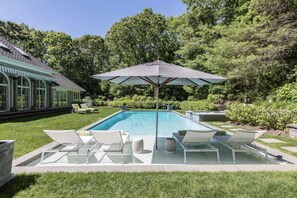 A swimming pool with lounge chairs and an umbrella, surrounded by greenery. - Bull Path Manor by Rove - 4BR Family Home With Pool & Hot Tub (East Hampton)