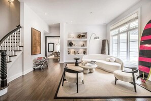 A modern living room with a staircase, a sofa, a coffee table, and a bookshelf. - Bull Path Manor by Rove - 4BR Family Home With Pool & Hot Tub (East Hampton)