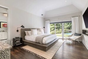A bedroom with a large bed, a desk, and a chair, a window with curtains, and a view of greenery outside. - Bull Path Manor by Rove - 4BR Family Home With Pool & Hot Tub (East Hampton)