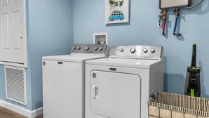 Un área de lavandería con dos lavadoras y secadoras Maytag, una secadora con conducto de ventilación, un calentador de agua y una cesta de ropa marcada con la etiqueta “LAUNDRY”. 