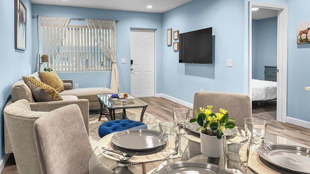Una sala moderna con comedor, paredes azul claro, un televisor de pantalla plana y una puerta visible que da a un dormitorio.